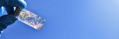 Gloved Hand Holding Glass Vial Of Colorful Microplastics Against Blue Sky